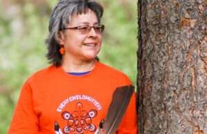 Native woman with shoulder length hair and glasses holding a feather and wearing an orange shirt.