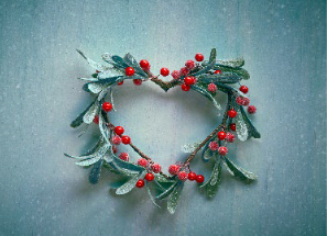 A heart-shaped wreath made of frosted evergreen leaves and red berries is set against a muted blue background.